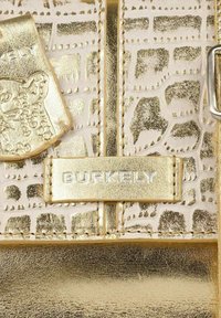 Bolso texturizado en dorado con un patrón de cocodrilo sintético, un acabado metálico y una etiqueta con el logo "BURKELY" en plata. Los acentos incluyen costuras cosidas.