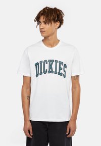 Vit t-shirt i bomull med ordet "DICKIES" i mörk teal i en bågformad text. Korta ärmar och rund halsdesign.