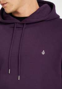 Lilla hoodie lavet af blødt stof, med en hætte med snøre, kengurulomme og et lille hvidt logo på brystet.