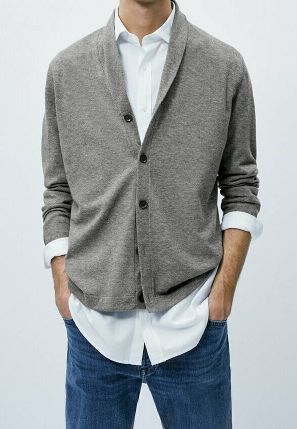 Homme portant un cardigan gris boutonné par-dessus une chemise blanche aux manches retroussées et un jean bleu, mains dans les poches.