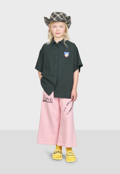 Barn klädd i en mörkgrön knäppskjorta med ett märke, rosa vida byxor med text och gula sandaler med bokstäver.