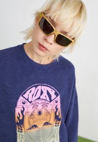 Sweatshirt bleu marine avec un grand graphique rose et orange portant le mot "ROXY". La personne porte des lunettes de soleil teintées en jaune avec une monture texturée.