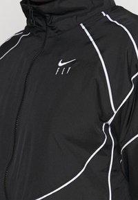 Chamarra deportiva negra con un alto cuello, líneas de acento blancas y un logo de Nike. Hecha de un material ligero y texturizado.