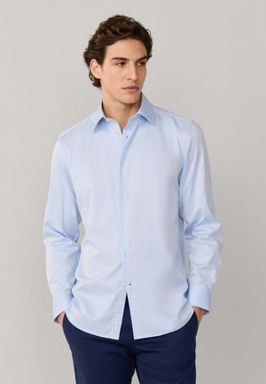 Jeune homme aux cheveux bouclés foncés portant une chemise bleu clair boutonnée et un pantalon bleu foncé, debout les mains dans les poches devant un fond neutre.