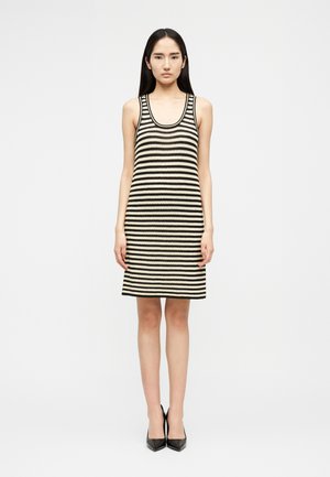 Νεαρή γυναίκα στέκεται με το πρόσωπο προς τα εμπρός φορώντας ένα αμάνικο, γόνατο μάκρος πλεκτό φόρεμα με ασπρόμαυρες ριγές και μαύρες γόβες με μυτερή μύτη.