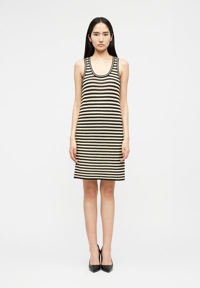 Jeune femme debout, face à l'avant, portant une robe tricotée sans manches, rayée noir et blanc, longueur au genou, et des escarpins noirs à bout pointu.