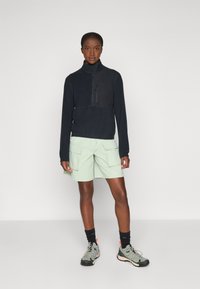 Schwarze Fleece-Zip-Jacke mit hohem Kragen, kombiniert mit hellgrünen Cargo-Shorts. Getragen mit schwarzen und grauen Sportschuhen und Socken.