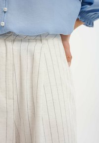 Himmelblaue Bluse mit perlweißen Knöpfen, kombiniert mit cremefarbenen, strukturierten Hosen mit vertikalen schwarzen Streifen und Seitentaschen.