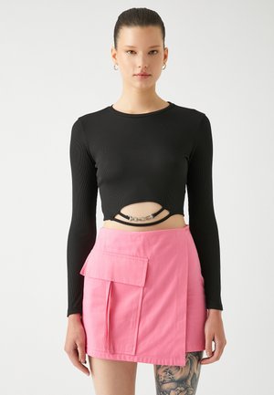 Zwart geribbeld langemouw crop top met een uitsnede en zilveren kettingaccent; gecombineerd met een roze mini-jurk met een voorzak en flap.