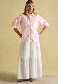 Svetloružová kockovaná košeľa s gombíkmi, doplnená dlhou bielou sukňou s výrezmi. Model má na sebe šedé balerínky.