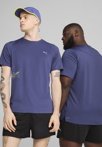 Violeta sporta krekli ar īsām piedurknēm, uz kojih ir Puma logotips un teksta detaļa, kopā ar melniem šortiem. Audums izskatās viegls un elpojošs.