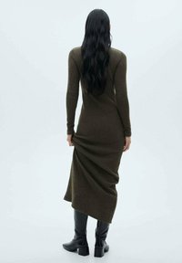 Robe longue et ajustée en tissu vert foncé à finition texturée, avec des manches longues et un ourlet droit. Portée avec des bottes noires jusqu'aux genoux.