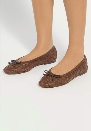 Füße, die braune gewebte Ballerinas mit kleinen Schleifen auf einem schlichten hellen Hintergrund tragen.