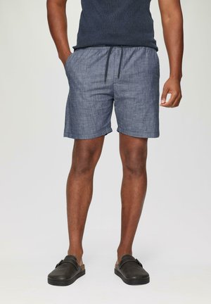 Blue chambray shorts with an elastic waistband and drawstring detail. Features a relaxed fit and rolled hem. Paired with black slip-on shoes.