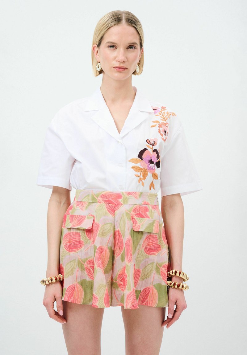 Camicetta bianca a maniche corte con ricamo floreale, abbinata a pantaloni corti rosa e verdi con motivo che presentano tasche laterali e una vestibilità comoda.