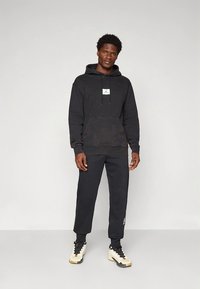 Čierna mikina s kapucňou, predným vreckom a šnúrkou, s malým logom. Kombinovaná s čiernymi teplákmi a čierno-bielymi teniskami.