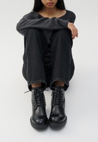 Stivali neri in pelle fino alla caviglia con punte rotonde, allacciatura frontale e suole texturizzate, abbinati a jeans scuri e un maglione grigio.