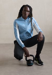 Modrá zipová bunda s bielymi akcentmi, čierne športové nohavice a čierne topánky. Drží čierny kettlebell s modrým podstavcom. Neutrálny pozadie.