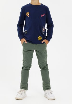 Dziecko ubrane w granatową bluzę z długim rękawem ozdobioną kolorowymi łatami, oliwkowozielone spodnie, białe trampki, stojące z rękami w kieszeniach.