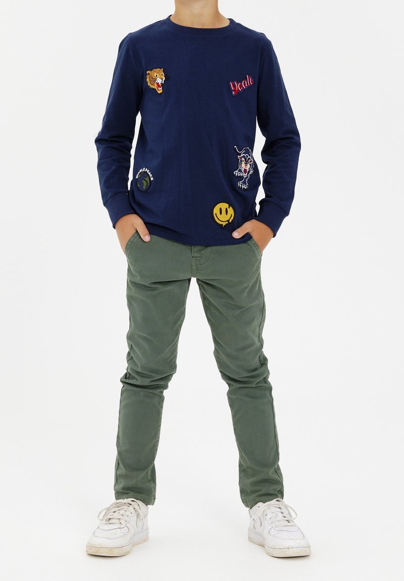 Dziecko ubrane w granatową bluzę z długim rękawem ozdobioną kolorowymi łatami, oliwkowozielone spodnie, białe trampki, stojące z rękami w kieszeniach.