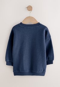 Tengerészkék pulóver kerek nyakkivágással és leejtett vállakkal. Puha anyag, bordázott mandzsettákkal és szegéllyel, egy fa vállfán lóg.