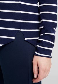 Personne portant une chemise à manches longues rayée bleu marine et blanche avec un détail d'ourlet queue de poisson, sur un pantalon bleu marine, main relâchée le long du corps.