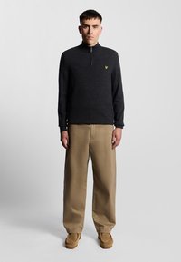 Mörkgrå zip-up-tröja med hög krage och en gul logotyp, matchad med lösa beiga byxor och beige skor. Enhetlig bakgrund.