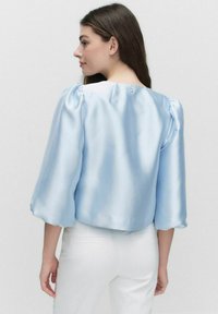 Ljusblå satinblus med puffärmar och rundad halsringning, med knappstängning bak. Slät textur och lätt lyster.