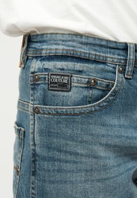 Κοντινό πλάνο μπλε τζιν με την ετικέτα Versace Jeans Couture στην μπροστινή τσέπη, φορεμένο με ένα λευκό πουκάμισο μασκαρισμένο μέσα.