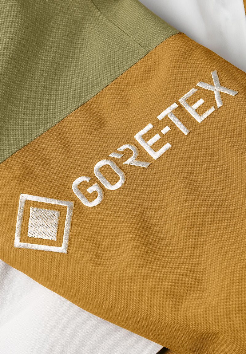 GORE-TEX-Patch auf einem Kleidungsstück mit olivgrünen und senfgelben Abschnitten, strukturiertem Gewebe, weißem gesticktem Logo und geometrischem Muster.