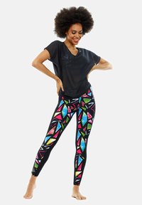 Schwarzes, transparentes T-Shirt kombiniert mit bunten, gemusterten Leggings, die geometrische Formen in Pink, Blau, Grün und Weiß auf einem schwarzen Hintergrund zeigen.