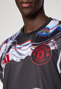 Maglietta atletica nera con motivo astratto colorato. Logo rosso del Manchester United e dettaglio a tre strisce Adidas sulla spalla.
