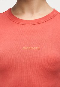 Gros plan sur un t-shirt à col rond côtelé de couleur corail orangé avec "element" brodé en fil assorti sur la poitrine.