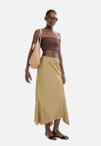 Kobieta w brązowej bluzce na ramiączkach, beżowej asymetrycznej spódnicy, czarnych sandałach na otwartym palcu, w dużych okularach, ze złotym mankietem na ramieniu i trzymająca plecioną torbę na ramię.