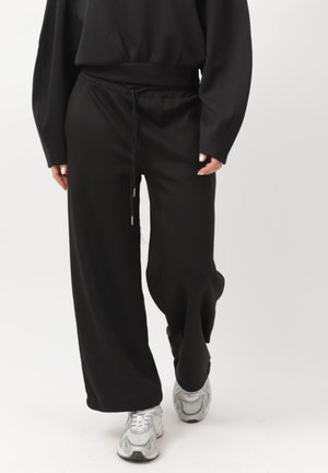 Osoba idąca, ubrana w luźne czarne spodnie z talią na sznurek, czarną bluzkę z długim rękawem i białe sneakersy na jednolitym białym tle.