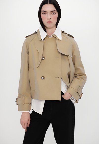 Trench coat crem cu croială scurtă, cu nasturi dubli, guler larg și manșete cu curea, purtat peste o cămașă albă și pantaloni negri.