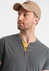 Šedá košile s krátkým rukávem a žlutou knoflíkovou lištou, doplněná béžovou čepicí s vyšívaným logem a texturovaným materiálem.