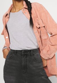 Camisa de corduroy rosa claro superpuesta sobre un suéter gris y una falda de mezclilla negra de cintura alta. Incluye botones, bolsillos y textura acanalada.