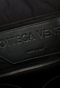 Étiquette en cuir noir Bottega Veneta avec nom de marque embossé et "Made in Italy" au-dessus d'une fermeture éclair métallique partiellement visible.