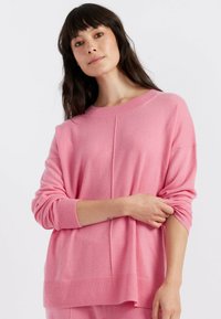 Pull en tricot rose avec une coupe décontractée, épaules tombantes, poignets et bas côtelés, présentant une texture subtile et des coutures visibles.