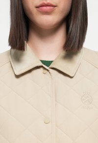 Beige quiltet jakke med kordfløjlkrave. Har knaplukning og et broderet logo på brystet. Grøn striktrøje under synlig.