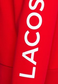 Červená mikina vyrobená z bavlněné směsi, s velkým bílým logem "LACOSTE" na rukávu, s pružným pasem a manžetami.