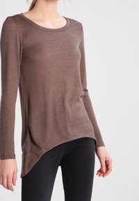 Braunfarbiges Langarmstrickoberteil mit Rundhalsausschnitt, weichem Material, asymmetrischem Saum und taillierter Passform, kombiniert mit dunklen Jeans.