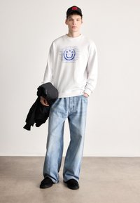 Λευκό φούτερ με μπλε γραφικό σχέδιο, φαρδιά ανοιχτό μπλε τζιν, μαύρα παπούτσια και μαύρο καπέλο, με το μπουφάν περασμένο στον αριστερό βραχίονα.