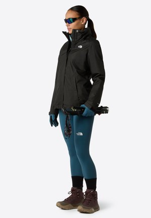 Kvinde i sort jakke, blå leggings, handsker og vandrestøvler, der holder foldede trekkingstænger, iført sports solbriller og står sidelæns.