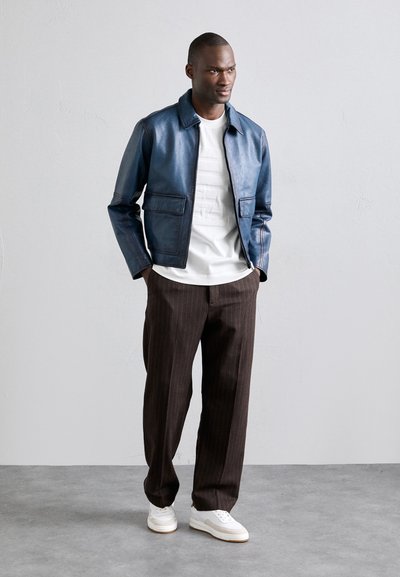 Veste en cuir marine avec deux poches avant, chemise blanche en dessous, pantalon marron à rayures, et baskets blanches. Texture lisse et coupe ajustée.