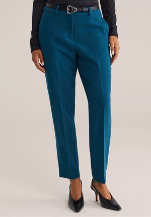 Femme portant un pantalon teal-tailleur avec plis sur le devant, ceinture noire, haut noir à manches longues et talons pointus noirs, debout sur un sol clair.