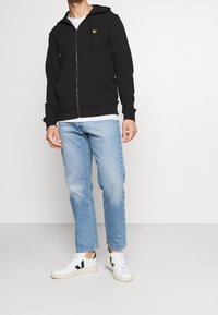 Černá mikina na zip s kapucí, předními kapsami a žlutým logem na hrudi, doplněná světle modrými džínami a bílými teniskami s černými detaily.