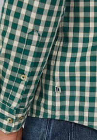 Groen en wit geruit lange mouwen overhemd manchet met knopen naast blauwe denim jeans en een klein stoffen merklabel.