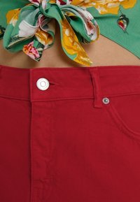 Pantaloni corti rossi in cotone a vita alta con chiusura con bottone frontale e tasca, abbinati a un papillon in tessuto floreale verde.